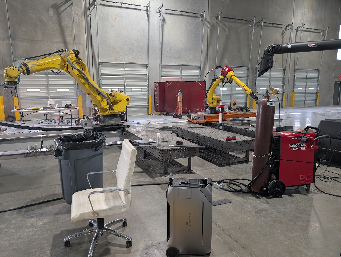 Wide view of the fabrication cell: FANUC robots, the perforated fixture table, welding equipment, and the surrounding cell infrastructure.